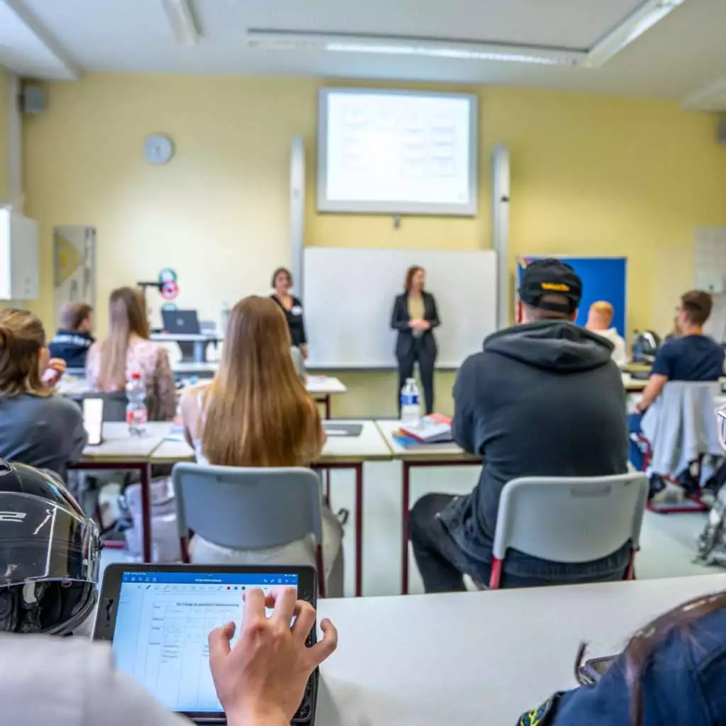 Mehrere Schüler und Schülerinnen sitzen an Tischen im Klassenzimmer. Vor der Tafel stehen zwei Lehrkräfte.